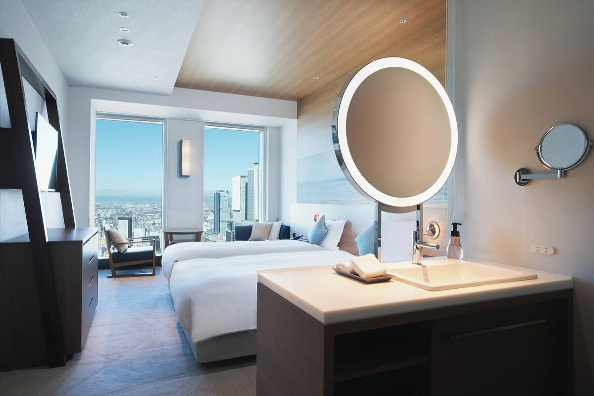 A modern hotel room with a large bed, a bedside table with a mirror, a desk, and a view of the city through large windows.