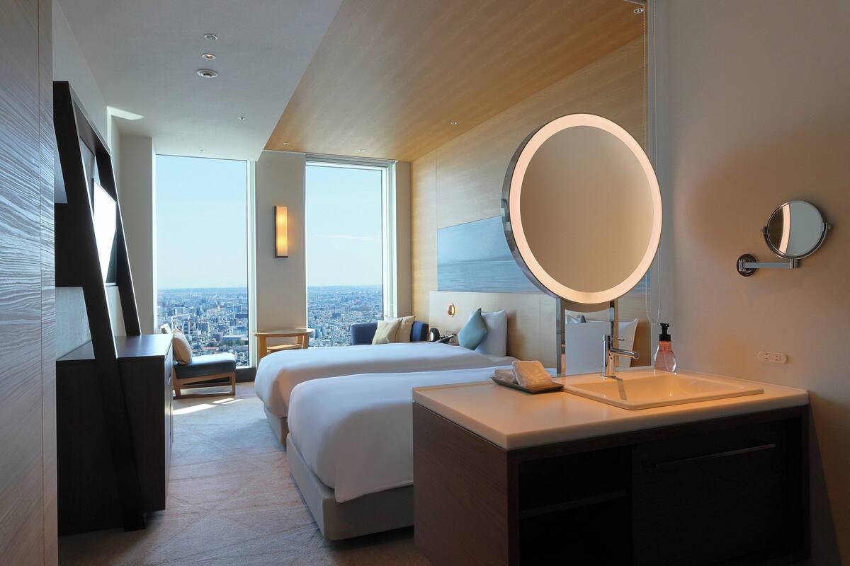 A modern hotel room with two beds, a large round mirror, a built-in sink, and a city view.