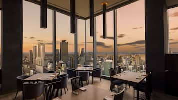A modern dining area with high ceilings and large windows offering a cityscape view.