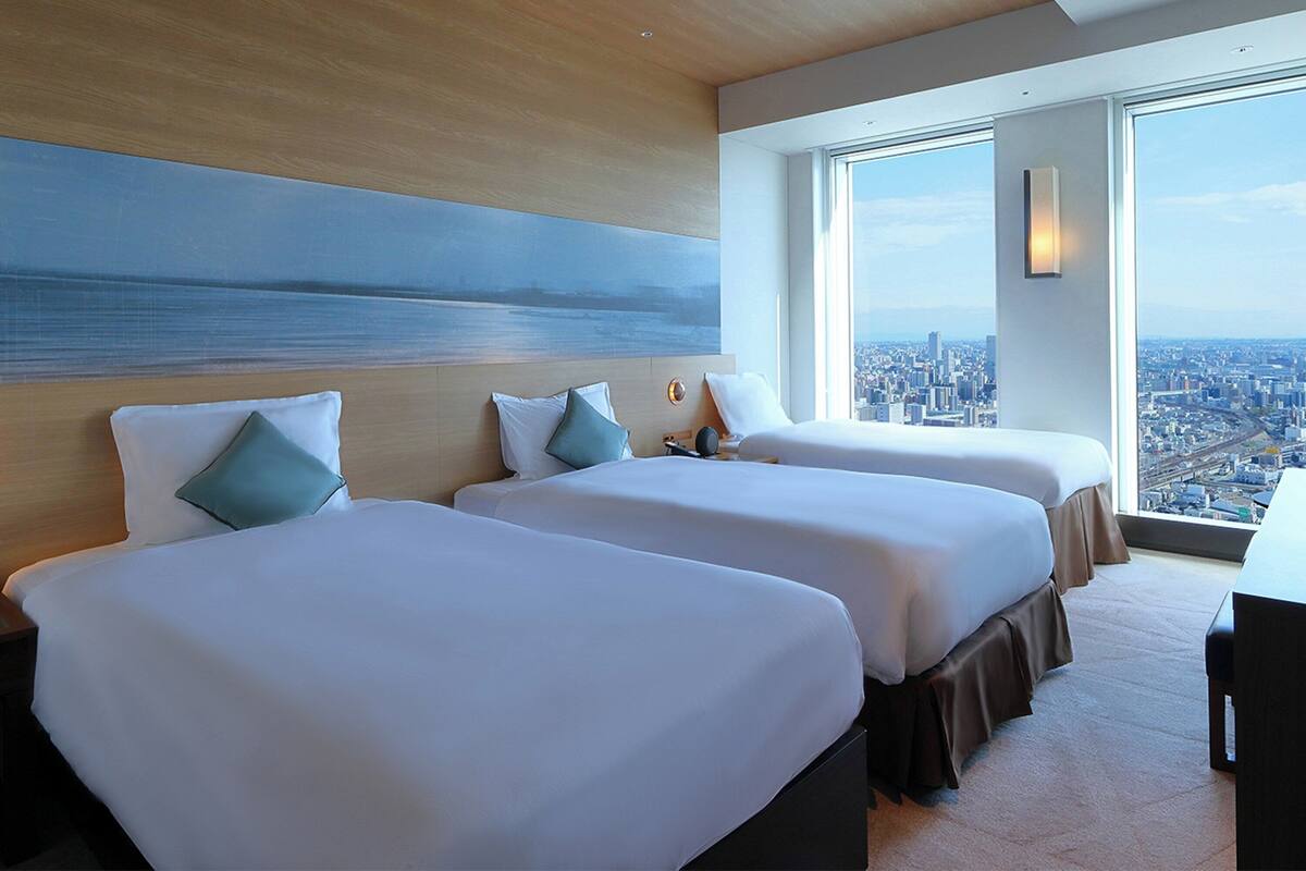A hotel room with two beds, a large window with a city view, and a painting on the wall.