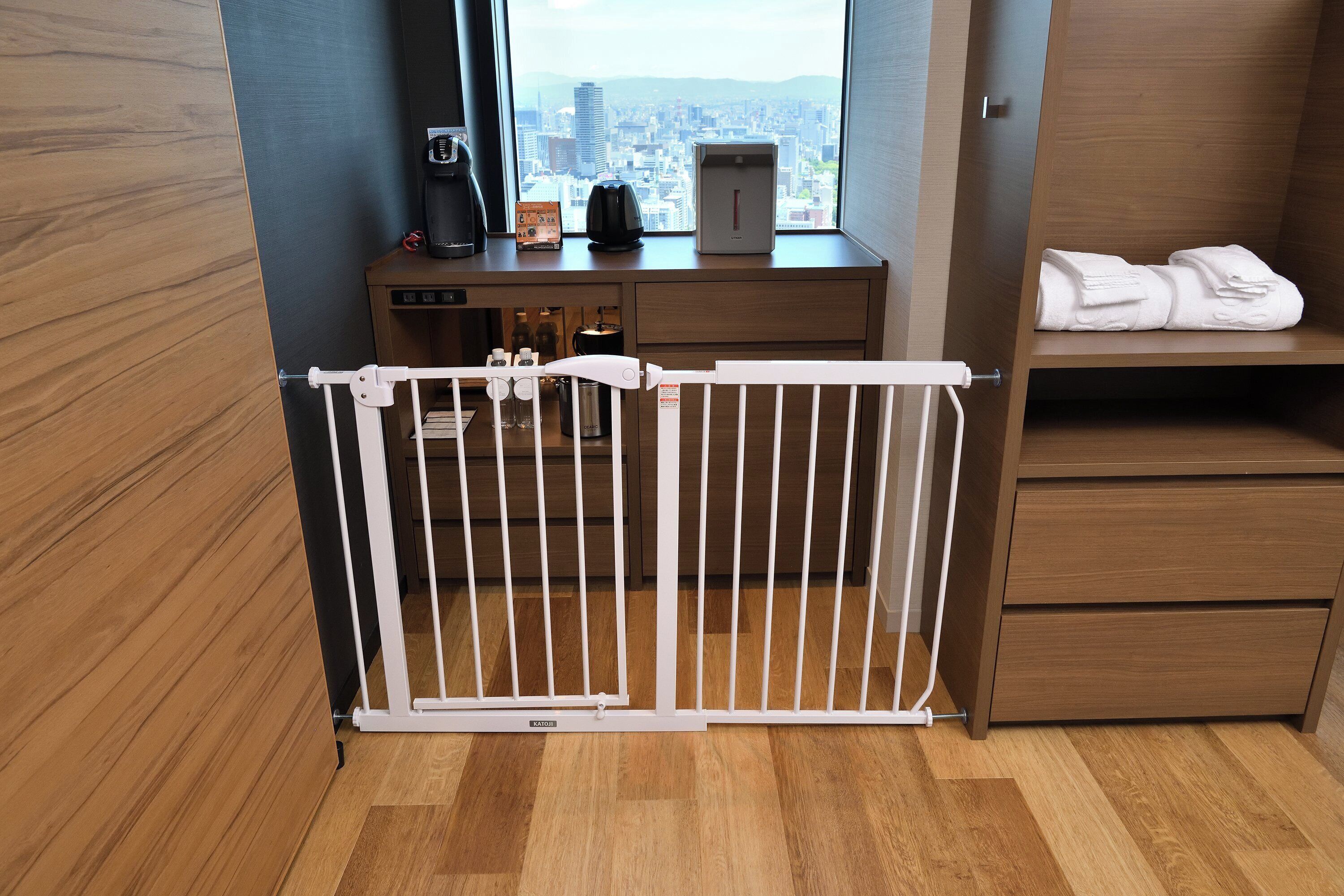 A hotel room with a view of a cityscape, a wooden desk with a coffee maker, a water dispenser, and neatly folded towels on a shelf.