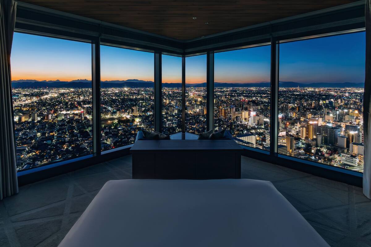 A high-rise hotel room with a large bed, a view of a cityscape at dusk, and a wooden ceiling.