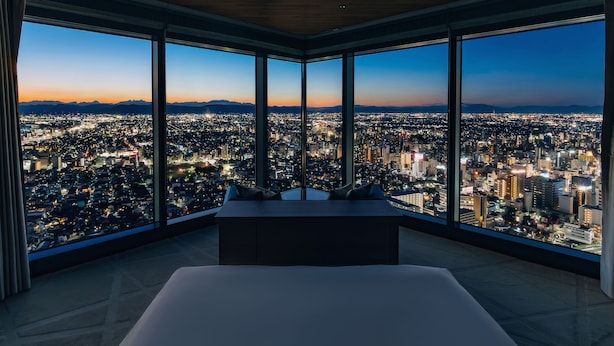 Une chambre d’hôtel située dans un immeuble de grande hauteur, avec un grand lit, une vue sur le paysage urbain au crépuscule et un plafond en bois.
