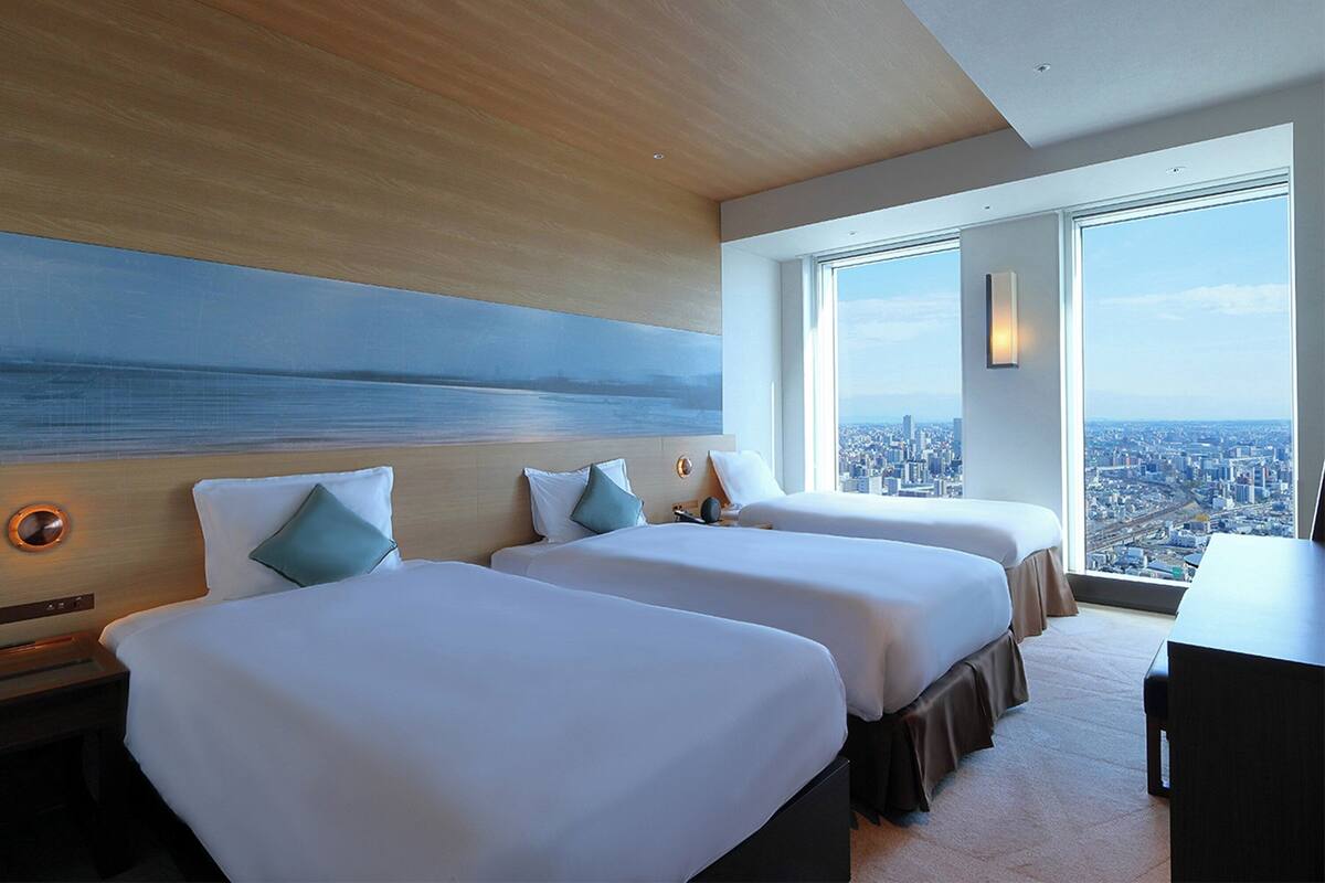 A hotel room with two beds, a desk, and a large window offering a city view.