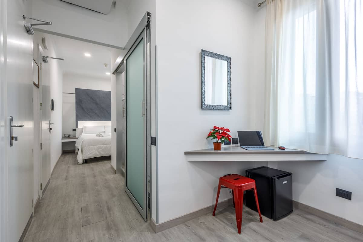 A modern hotel room with a bed, a desk with a laptop, a small red stool, and a compact black refrigerator.