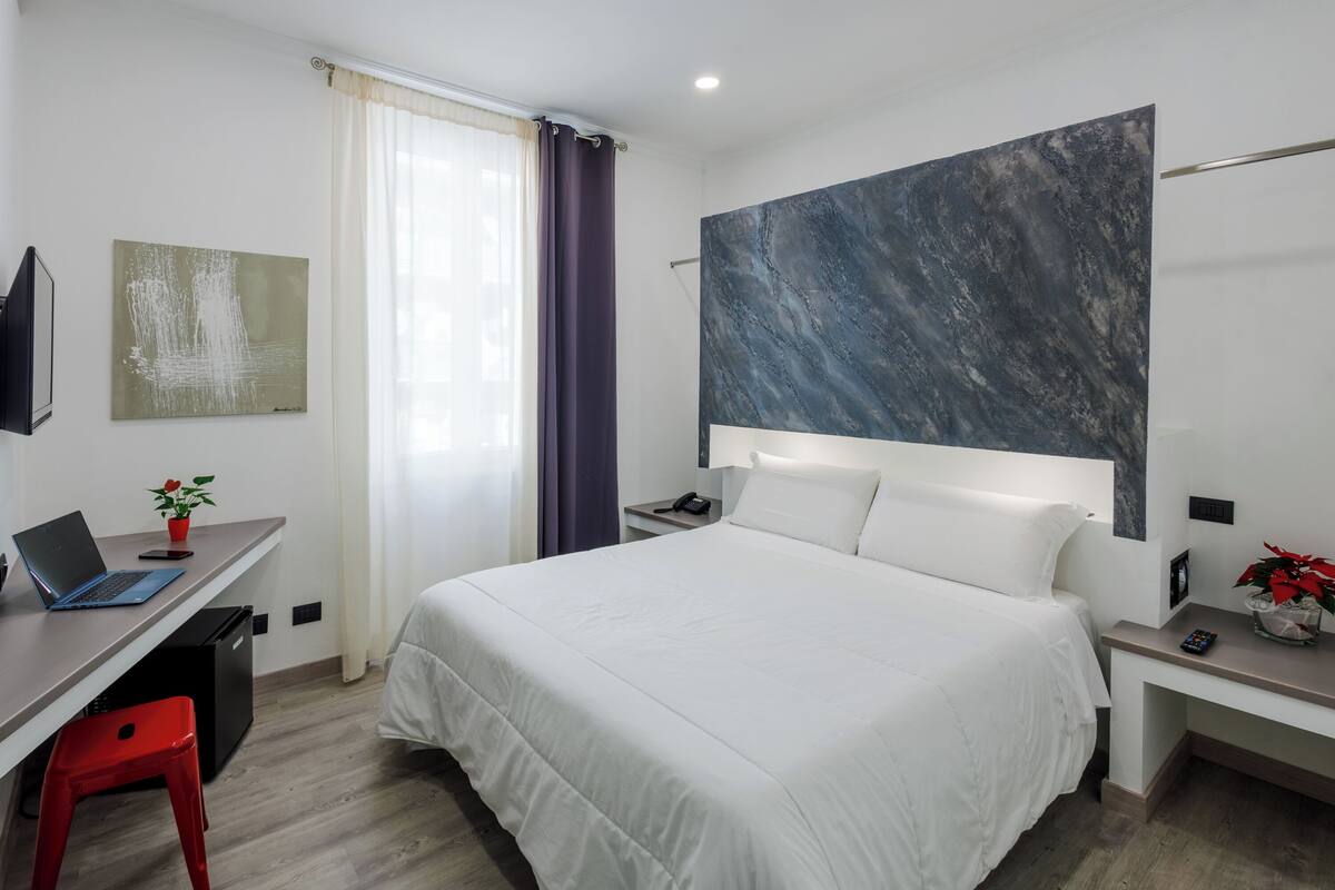 A modern hotel room with a large bed, a desk with a laptop, a red stool, a nightstand with a phone, and a large abstract painting on the wall.
