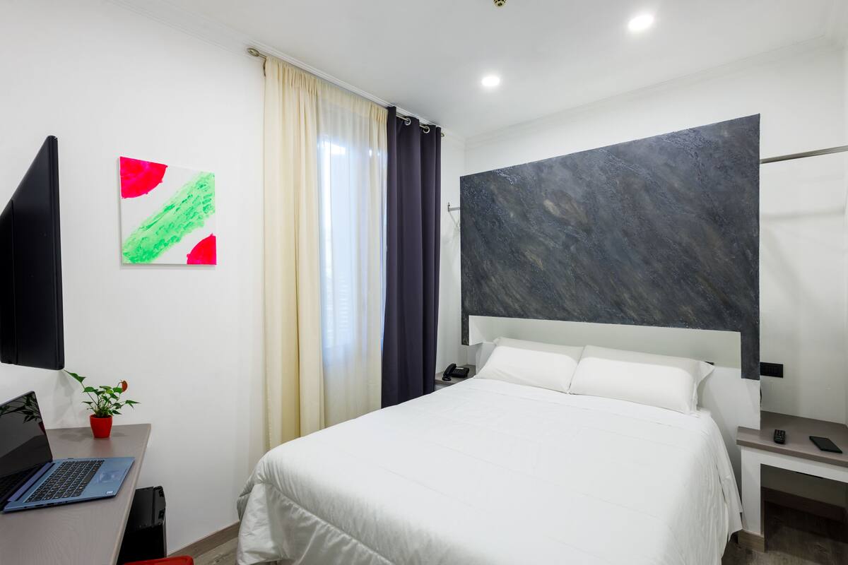 A modern hotel room with a bed, a desk with a laptop, a potted plant, and a large abstract painting on the wall.