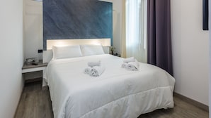 Een moderne hotelkamer met een groot bed, witte lakens, een nachtkastje, een afstandsbediening en een kunstwerk aan de muur.