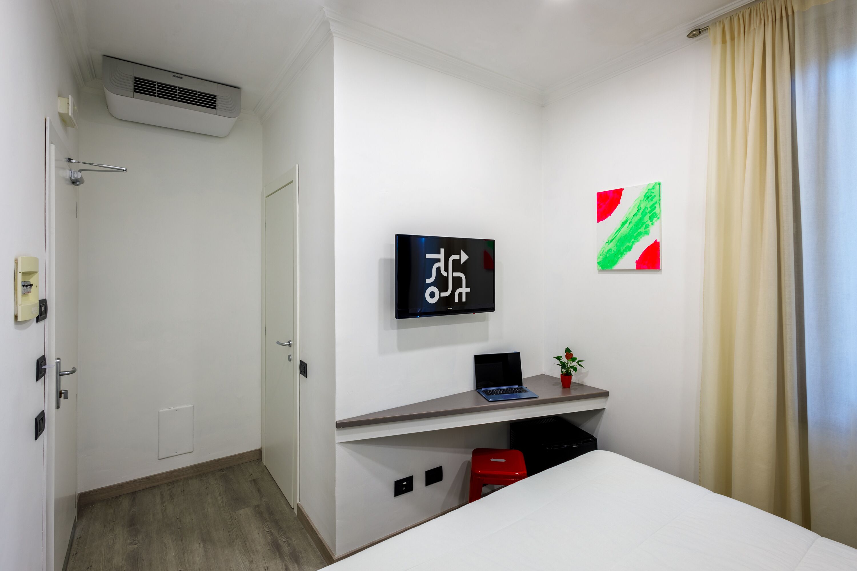 Une chambre d’hôtel avec un lit, un bureau avec un ordinateur portable, une petite plante, une télévision et une porte sur laquelle est fixé un porte-serviettes.