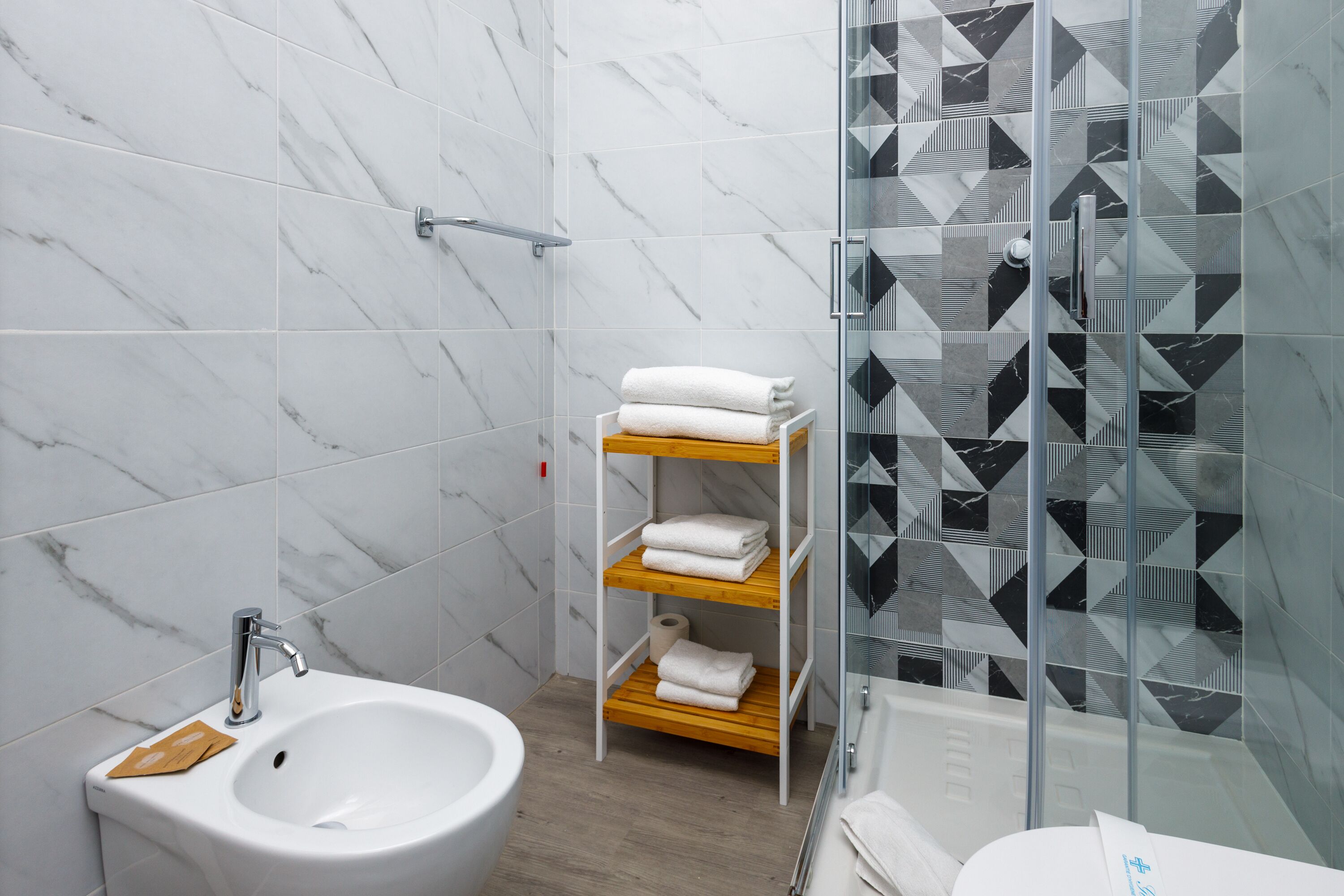 A modern bathroom with a white sink, marble tiles, a glass shower enclosure, and a wooden shelf with towels.