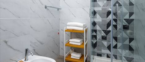 A modern bathroom with a white sink, marble tiles, a glass shower enclosure, and a wooden shelf with towels.
