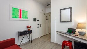 Een moderne hotelkamer met een houten vloer, een rode bank, een klein rode kruk, een bureau met een laptop, een spiegel en een abstract schilderij in groen en rood.