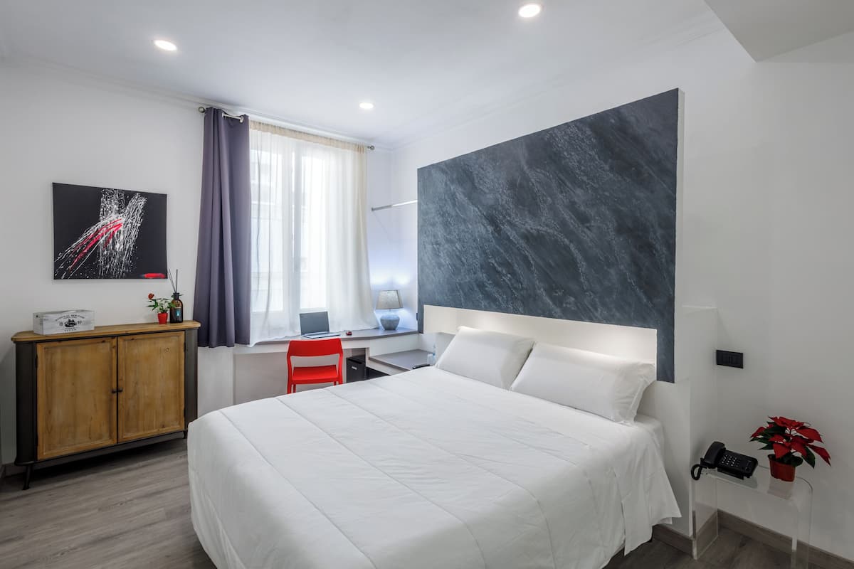 A modern hotel room with a large bed, a desk with a chair, a wardrobe, and a large abstract painting on the wall.