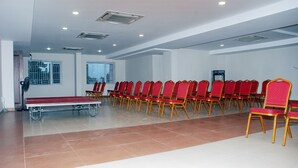 Un salón de conferencias con filas de sillas rojas, una mesa y una pantalla de proyector.