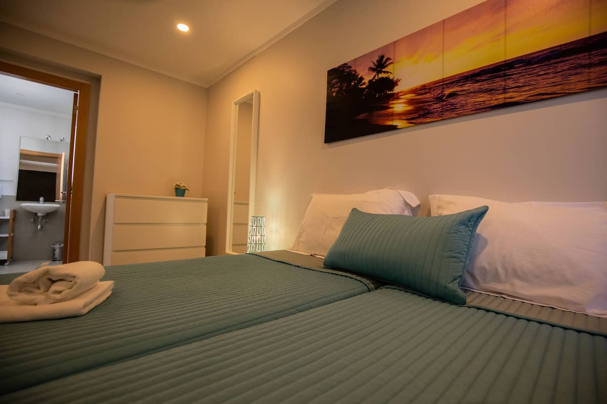 A hotel room with a bed, pillows, a nightstand, a wall-mounted painting, and a bathroom visible through an open door.
