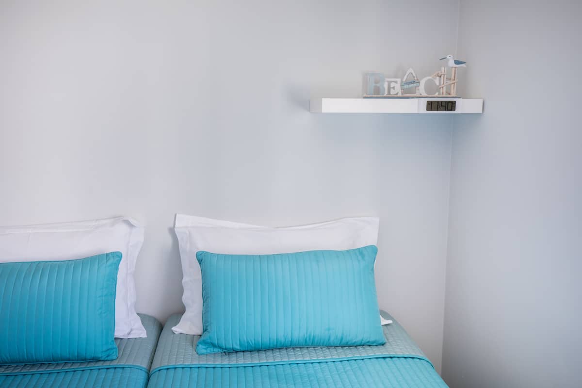 Two beds with blue bedding and white pillows, a shelf with decorative items, and a digital clock.