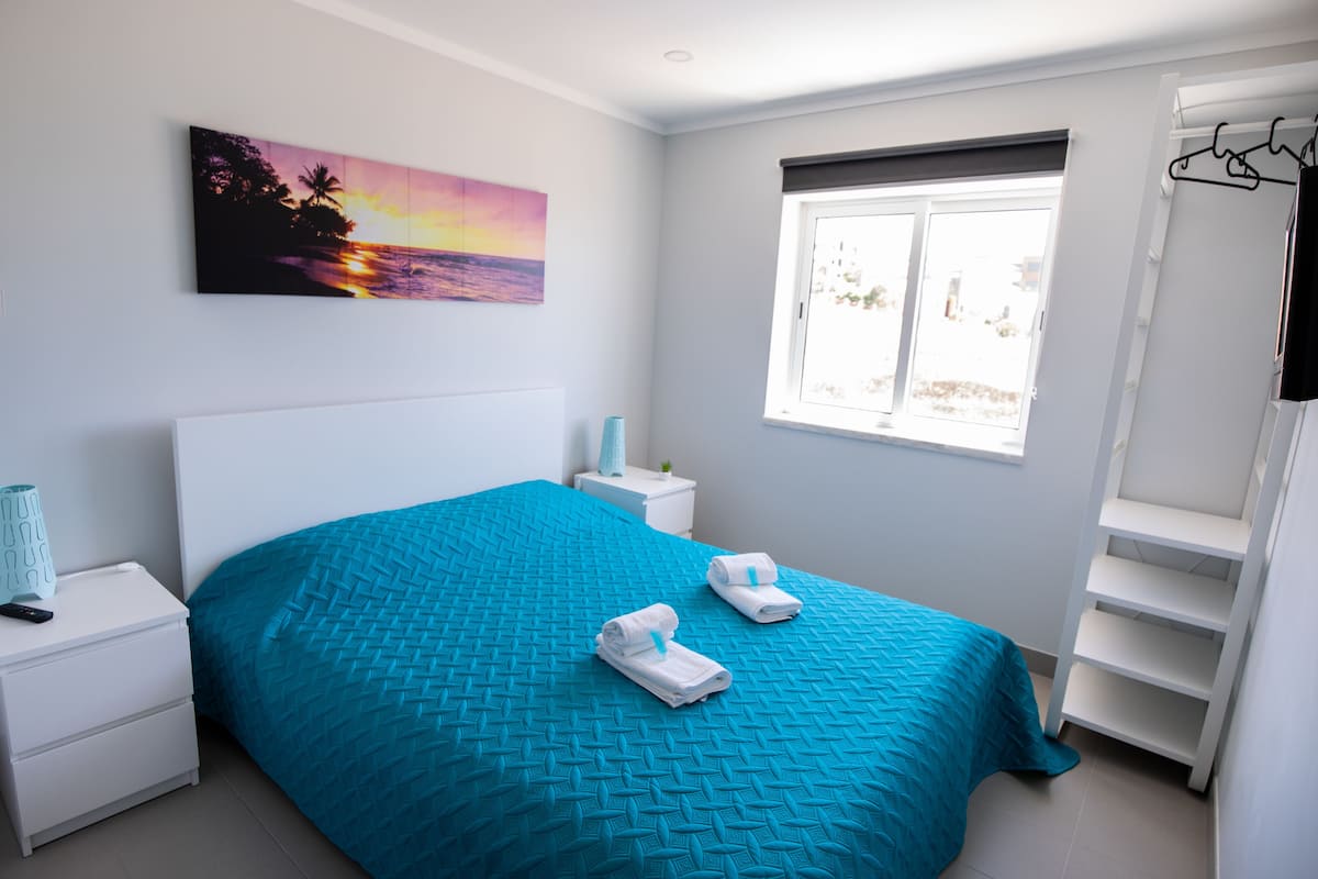A bedroom with a bed, a nightstand, a bedside table, a window, and a wall-mounted shelf.