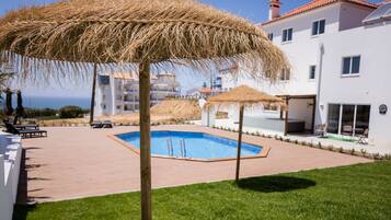 Una piscina rodeada por una terraza de madera, con un paraguas de paja y un edificio blanco al fondo.
