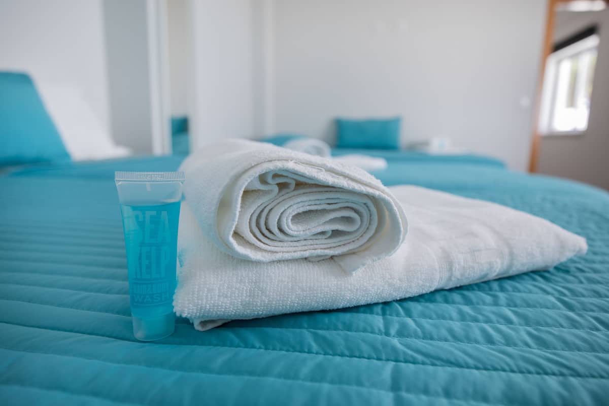 A neatly made bed with a blue quilt, a rolled white towel, and a blue bottle of body wash on the side.