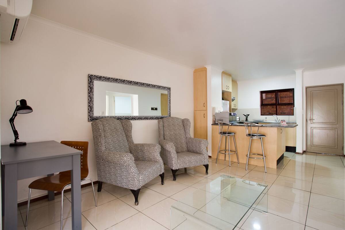 A modern living room with two leopard print armchairs, a small table with a lamp, and a glass coffee table. A kitchen area with a bar stool and a refrigerator is visible in the background.