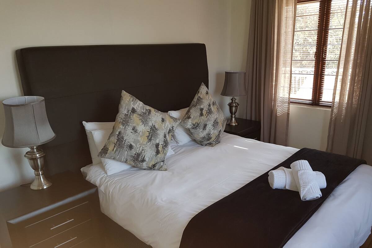A neatly made bed with white linens, two patterned pillows, a dark headboard, a bedside lamp, and a window with curtains.