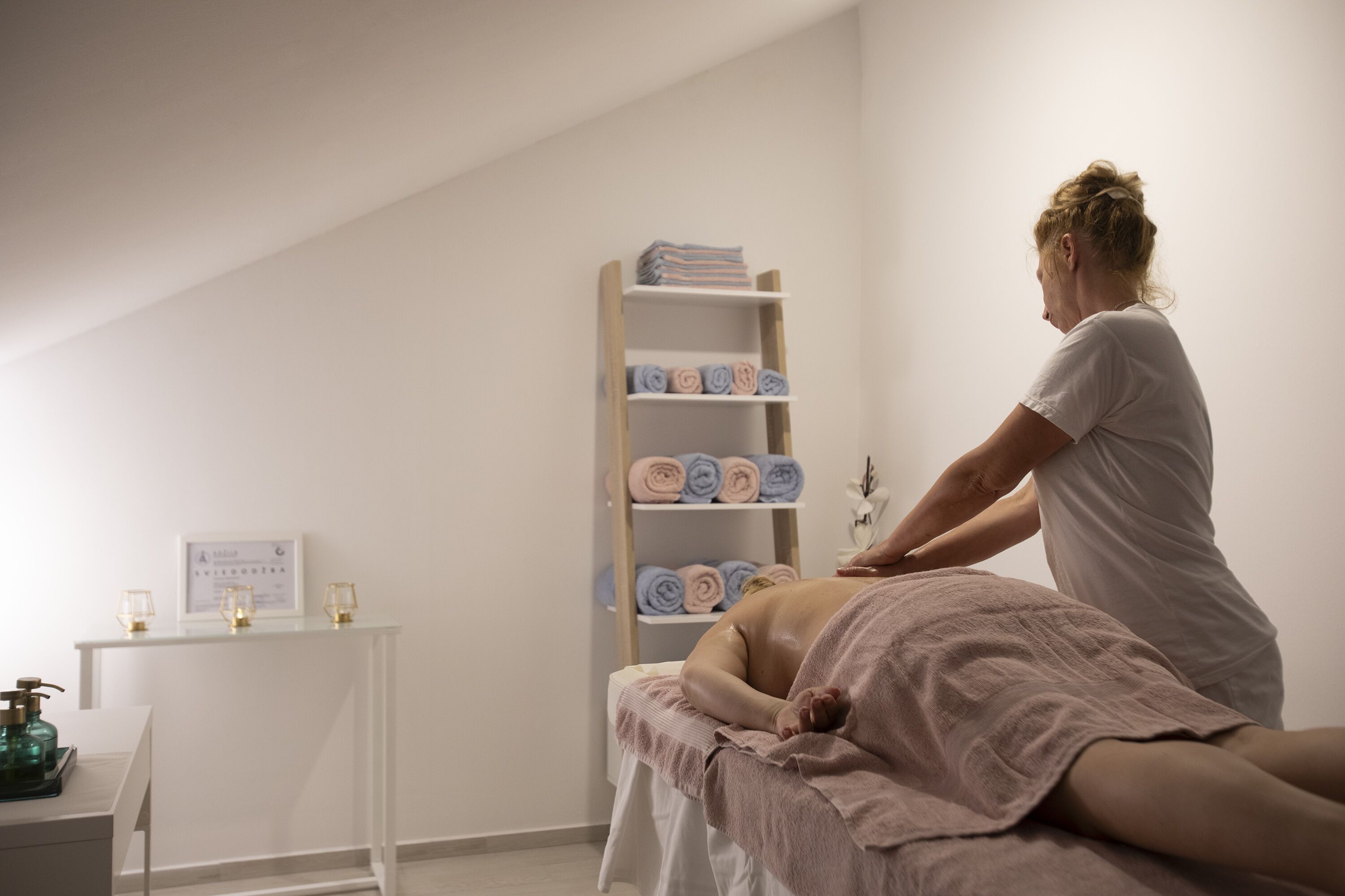 A massage therapy room with a massage table, a shelf with towels, a small table with a bottle and glasses, and a framed certificate on the wall.