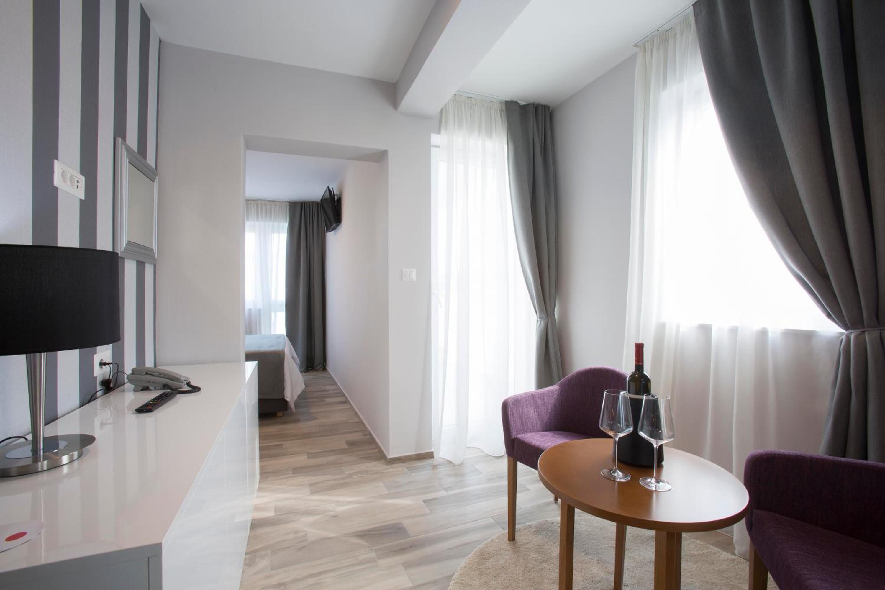 Quarto de hotel moderno com escrivaninha branca, sofá roxo, mesa de madeira com copos de vinho e uma cama ao fundo.
