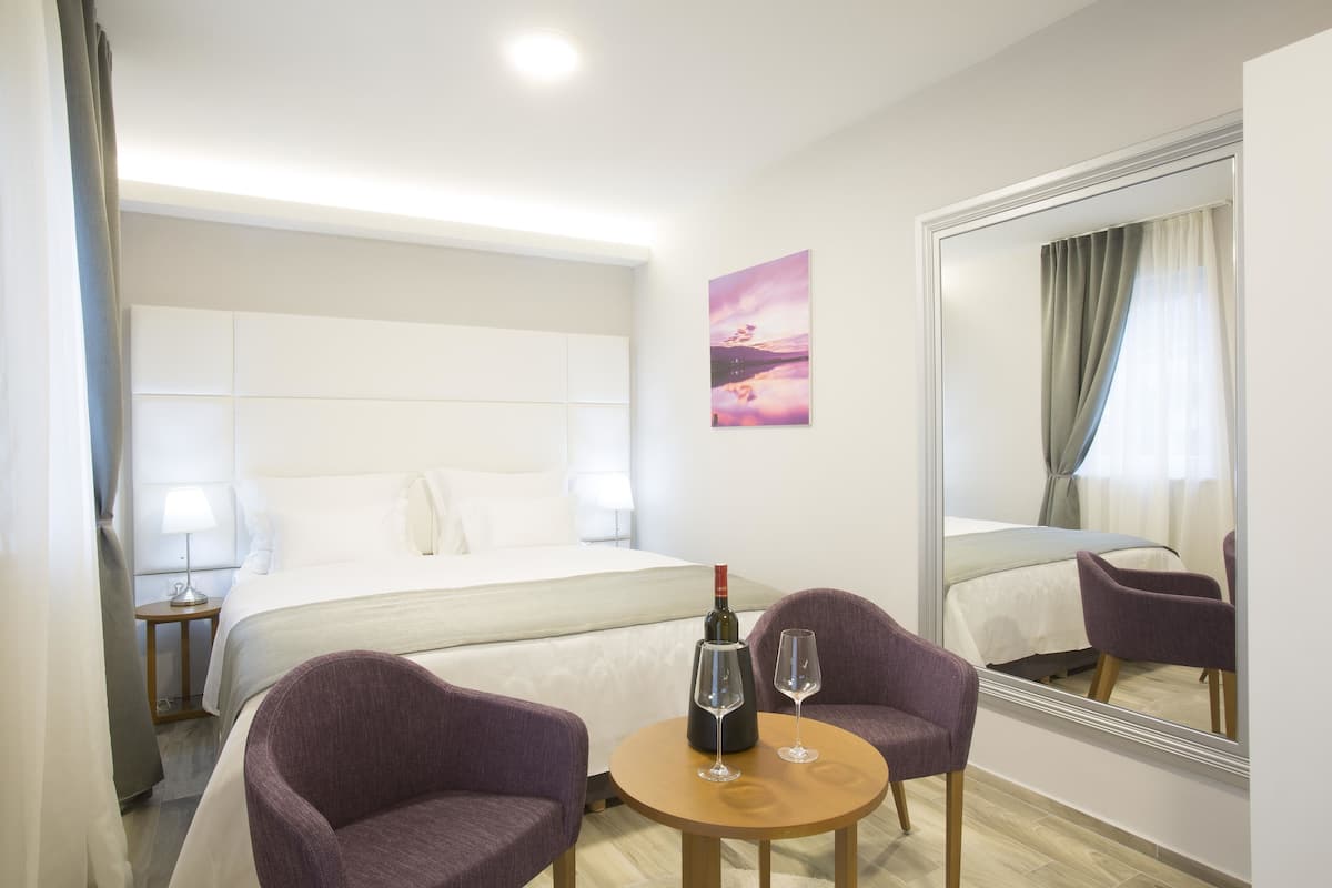 A hotel room with a bed, two purple armchairs, a small round table with a bottle and two glasses, a nightstand with a lamp, and a large mirror reflecting the room.