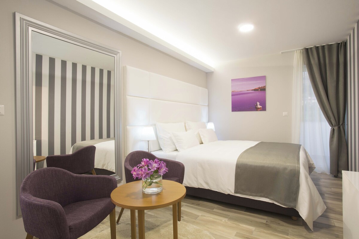 Habitación de hotel con una cama, una silla, una mesa con un jarrón de flores, un cuadro en la pared y una ventana con cortinas.