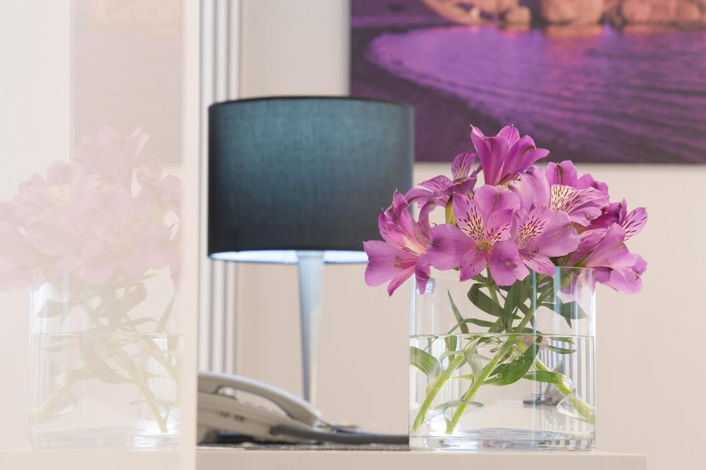 Eine klare Glasvase mit lila Blumen auf einem Tisch, eine schwarze Lampe und ein Telefon im Hintergrund.