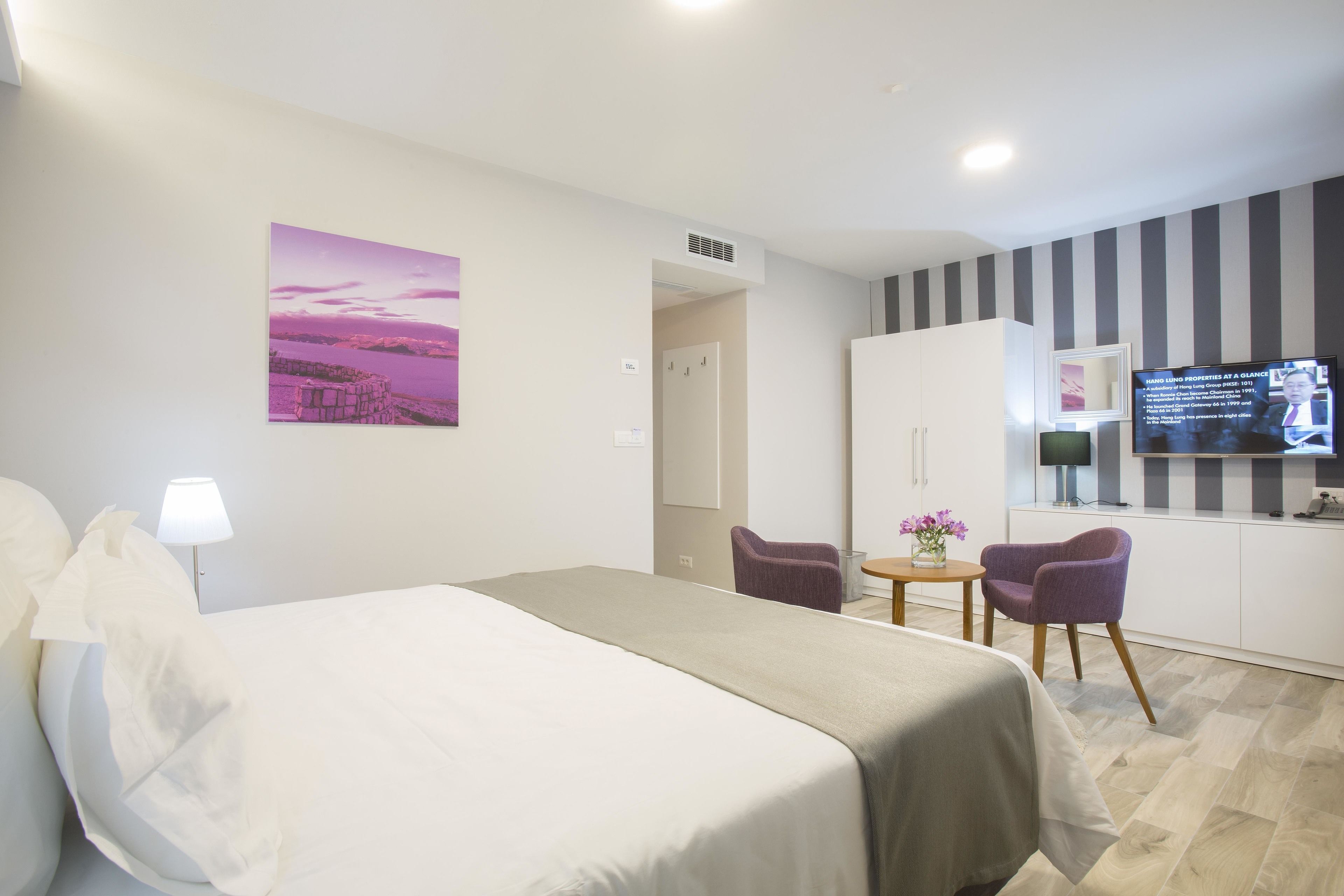 A hotel room with a bed, a desk, a chair, a TV, and a painting on the wall.