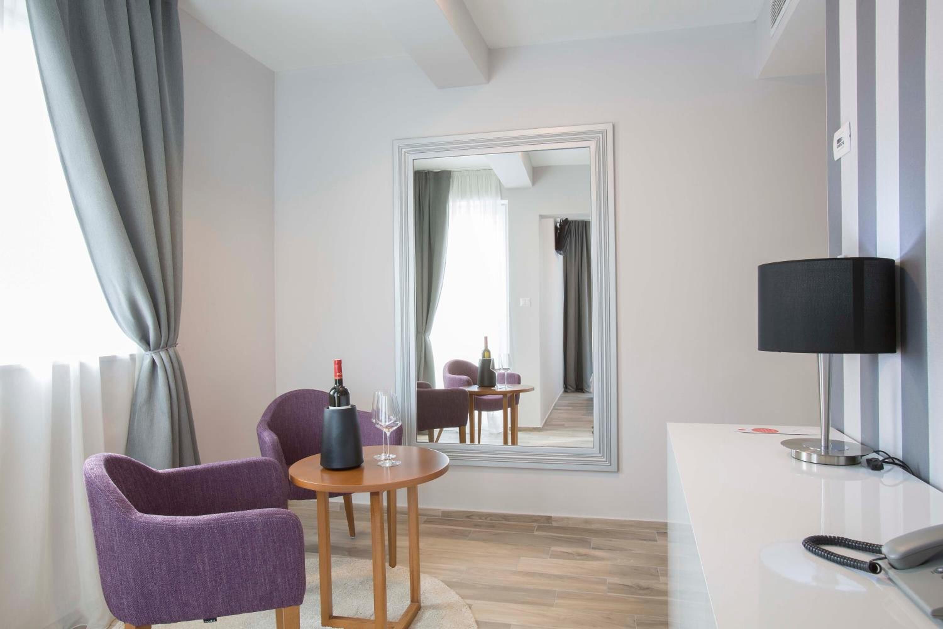 A modern hotel room with a large mirror, a wooden table with a wine bottle and glass, purple chairs, a desk with a lamp, and a telephone.