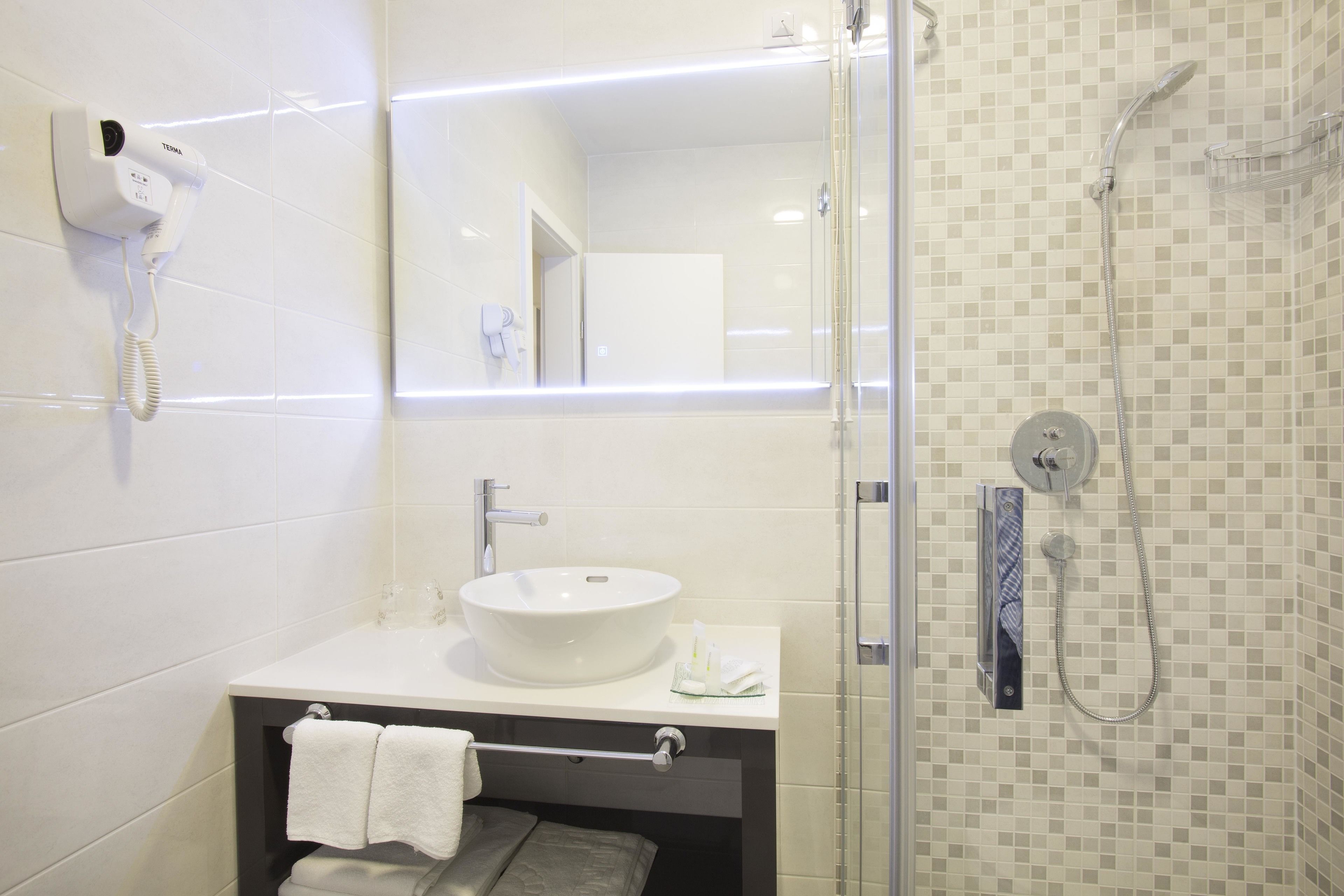 Une salle de bain avec un lavabo blanc, un porte-serviettes, un sèche-cheveux et une douche dont le mur est recouvert de carreaux.