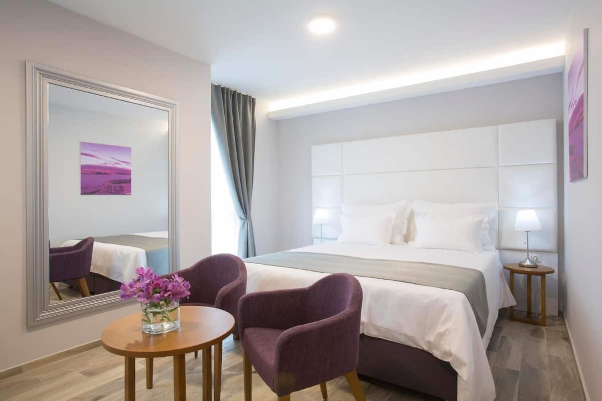 A hotel room with a large bed, two purple armchairs, a round wooden table with a vase of flowers, a large mirror, and a view of the cityscape.