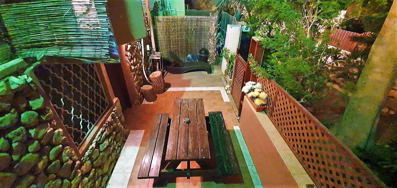 A patio area with a wooden table and benches, surrounded by a bamboo fence and greenery.