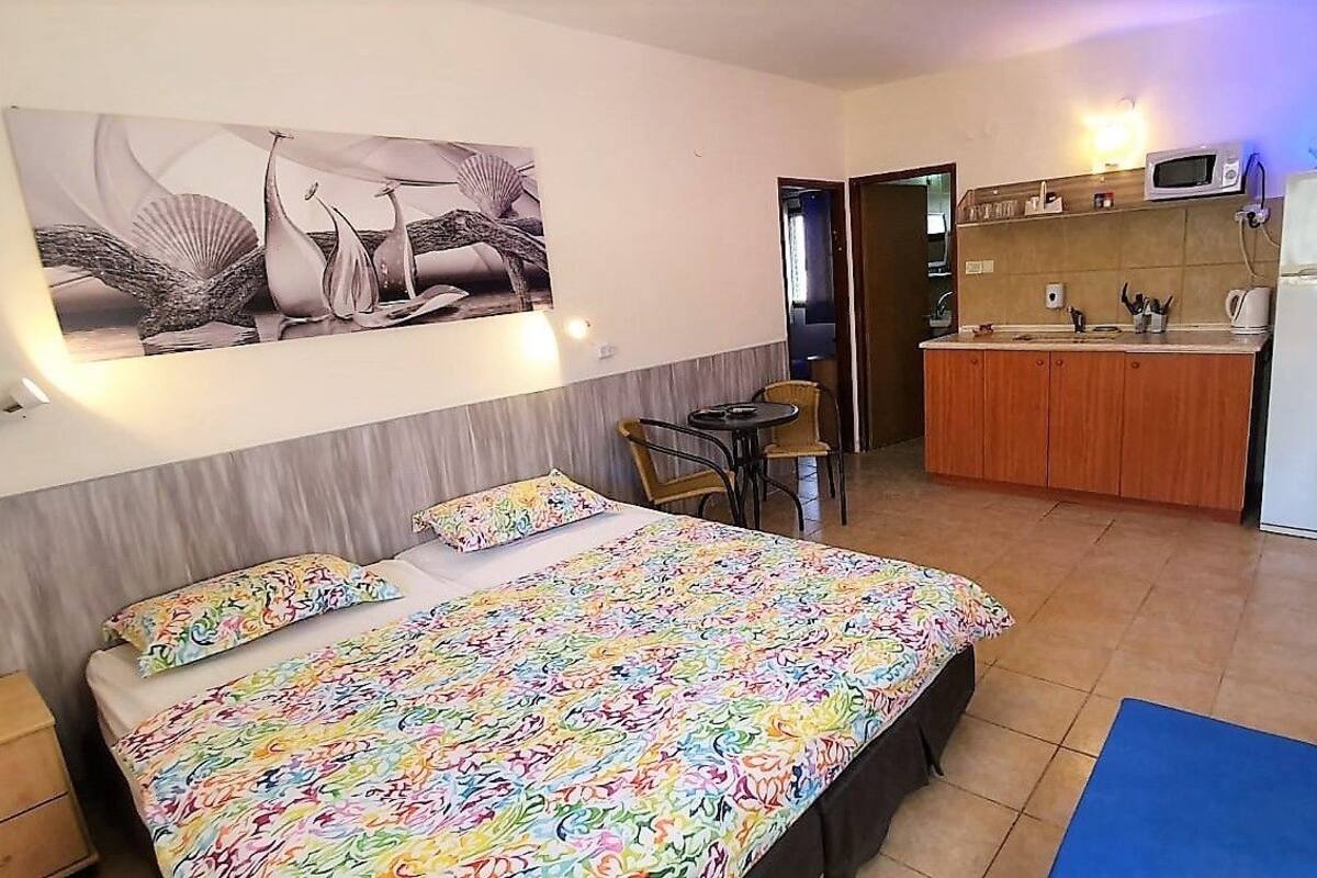 A bedroom with a bed, a small table, and a chair. There is a kitchenette with a microwave and a refrigerator.