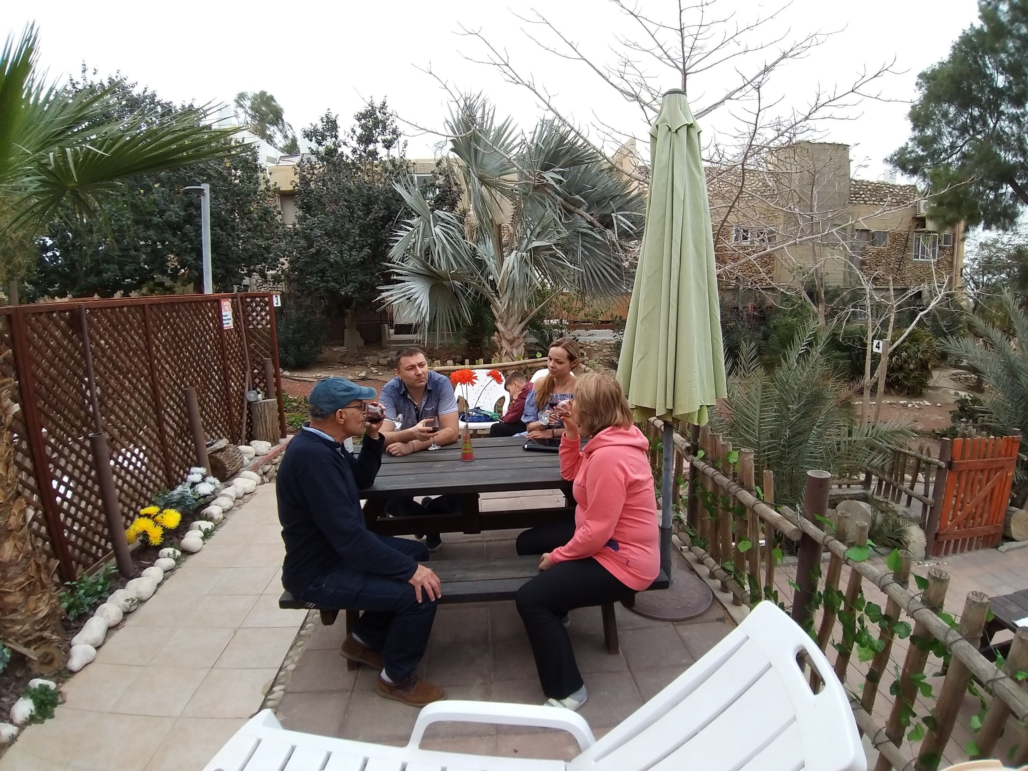 Un grupo de personas sentadas alrededor de una mesa en un patio al aire libre con valla de madera, plantas y un edificio de fondo.