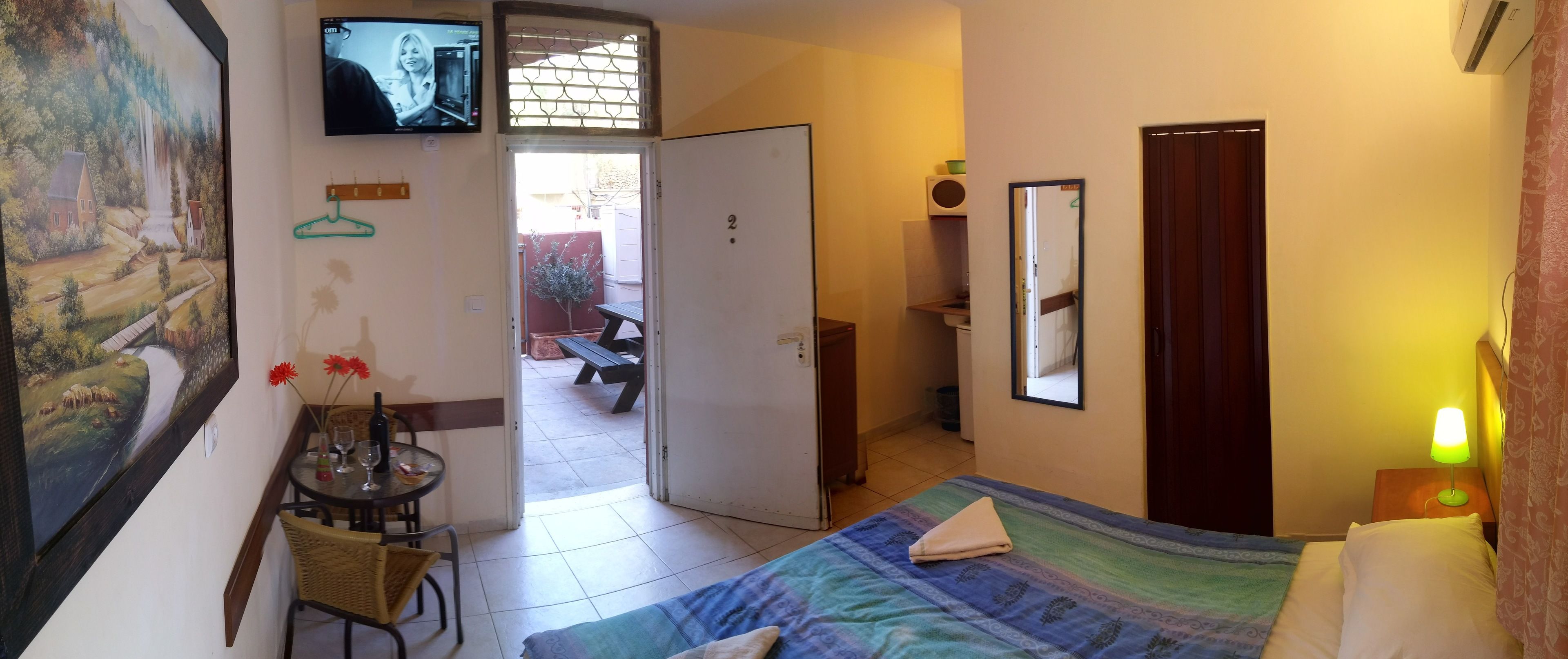 A hotel room with a bed, a TV, a dining table with chairs, a small table with a bottle and glasses, a bench outside, and a door leading to a patio.