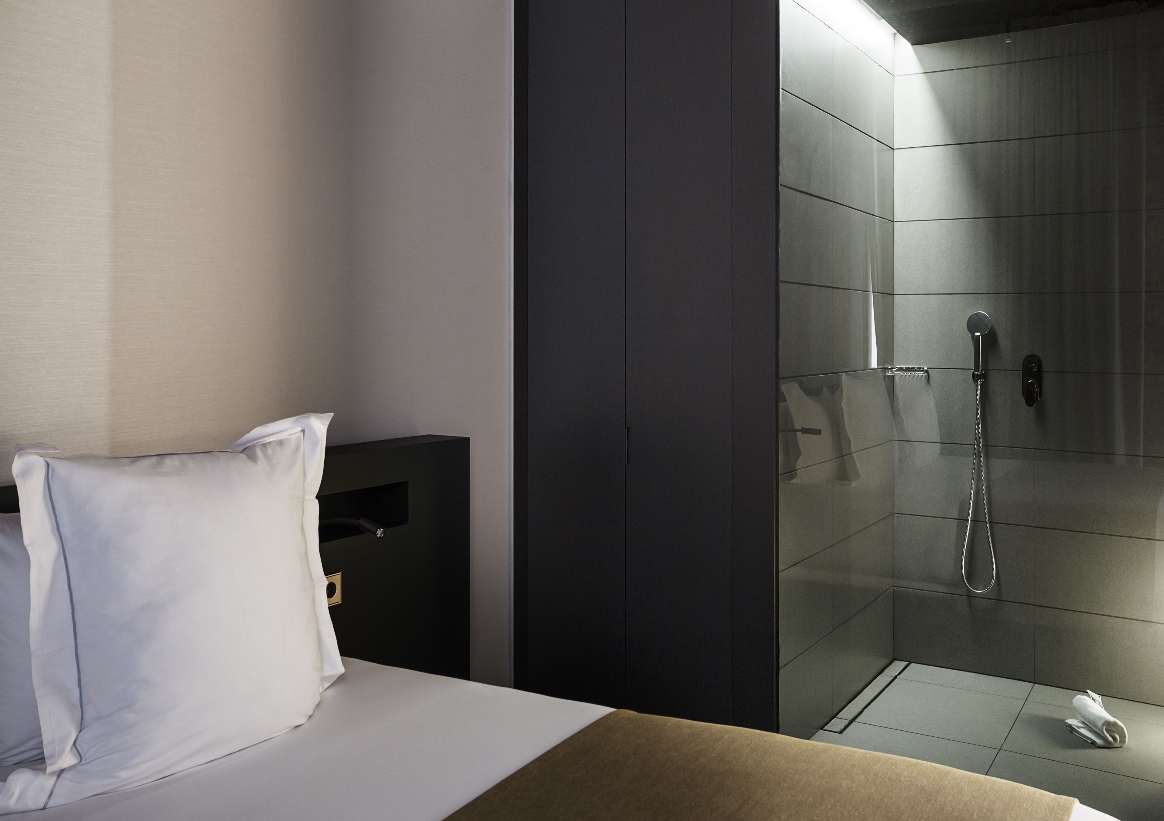 Una habitación de hotel con cama, mesita de noche, armario y zona de ducha con mampara de vidrio.