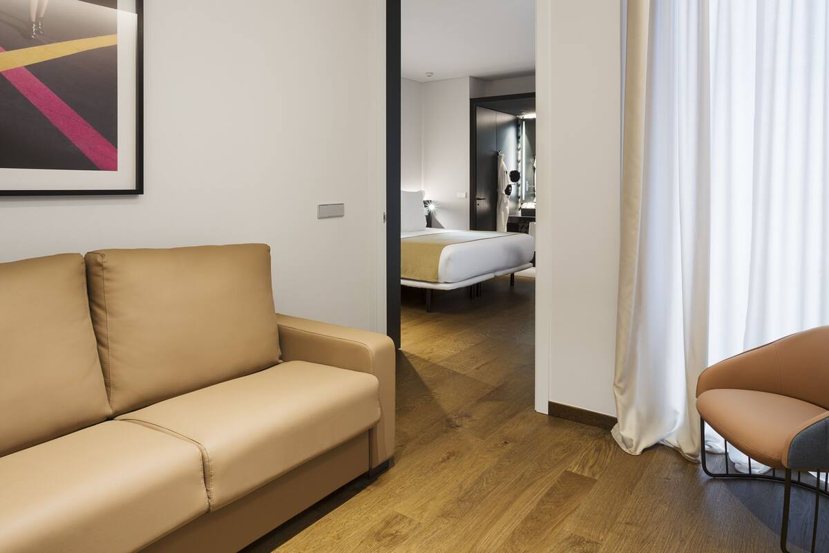 A hotel room with a beige sofa, a bed with a white mattress, a wooden floor, and a framed picture on the wall.