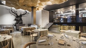 A modern restaurant interior with geometric wooden ceiling panels, a large tree mural, and a wine cellar visible through a glass wall. - One Shot Mercat (Valencia)