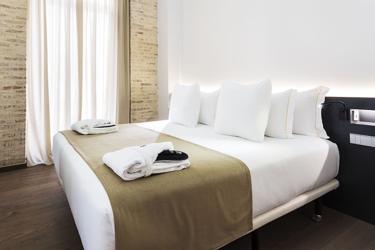 A neatly made bed with white linens and a brown throw blanket, a set of folded towels, and a black cap on the bed.