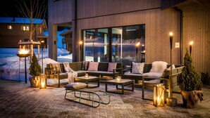 An outdoor seating area with sofas, tables, and lanterns, set against a modern building with large windows. - LA SOA Chalets & Eventlodge (Schattwald)