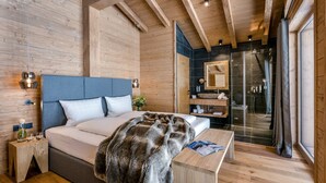 A bedroom with a wooden ceiling, a large bed, a bedside table, a nightstand, a mirror, a bathroom with a glass shower, and a window. - LA SOA Chalets & Eventlodge (Schattwald)