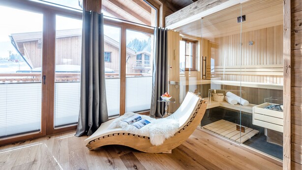 A wooden cabin with a glass-enclosed sauna, a reclining chair with a fur throw, and a small table with a bowl of fruit. - LA SOA Chalets & Eventlodge (Schattwald)