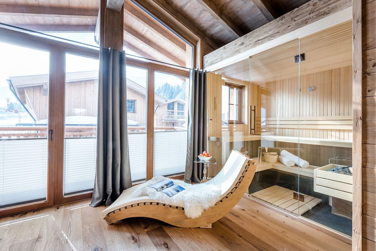 Una cabaña de madera con sauna de vidrio, una silla reclinable con manta de piel y una mesa pequeña con un cuenco de fruta.