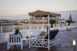 Una terrazza sul tetto con un'area salotto coperta, sedie bianche e un tavolo con un vaso di fiori.