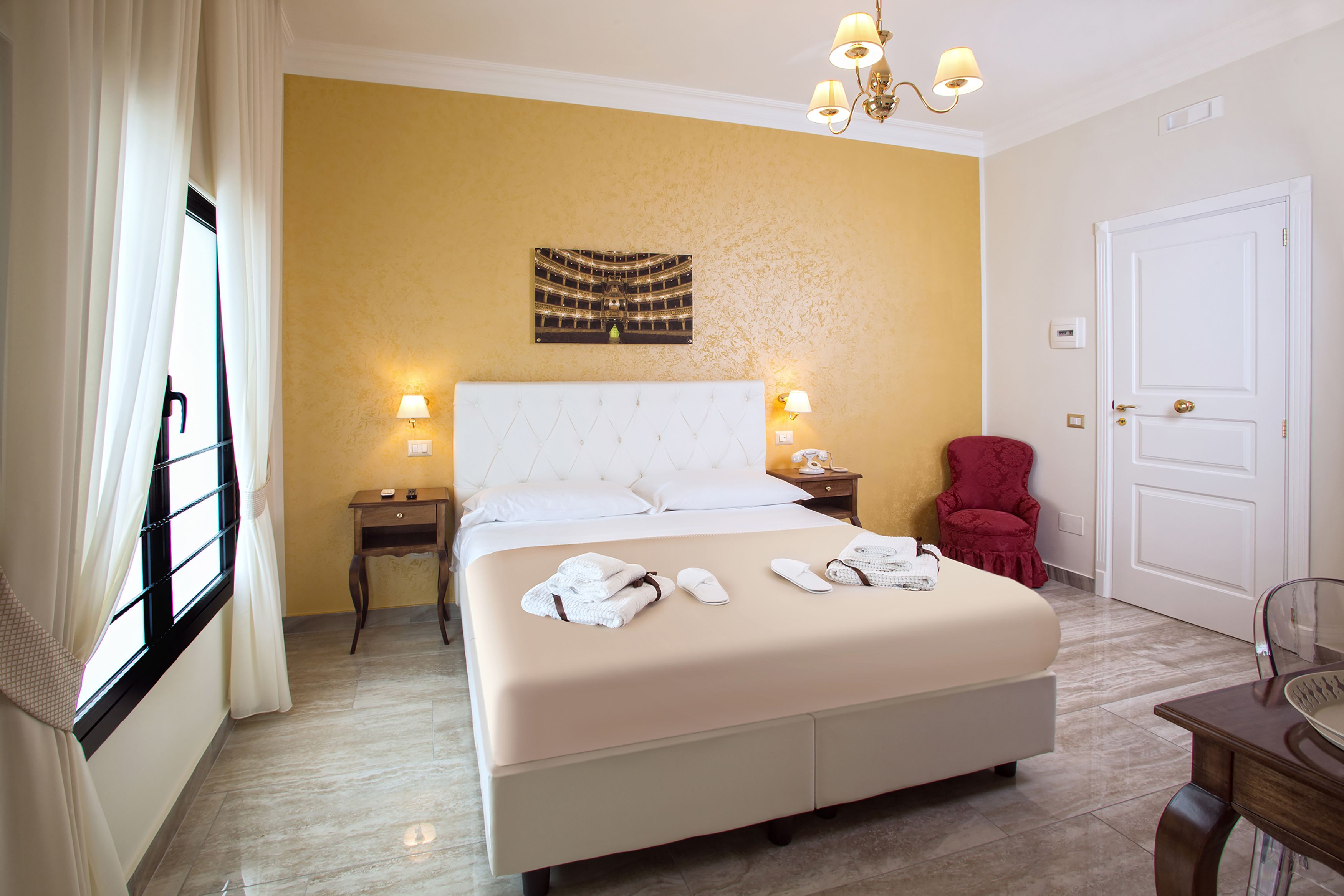 A hotel room with a large bed, two bedside tables, a red chair, a chandelier, and a door.