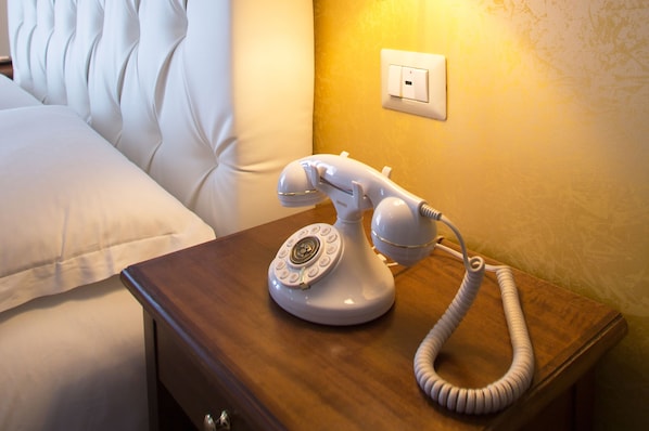 A white rotary phone on a wooden bedside table next to a bed with white bedding. - NeapolitanTrips Hotel (Naples)