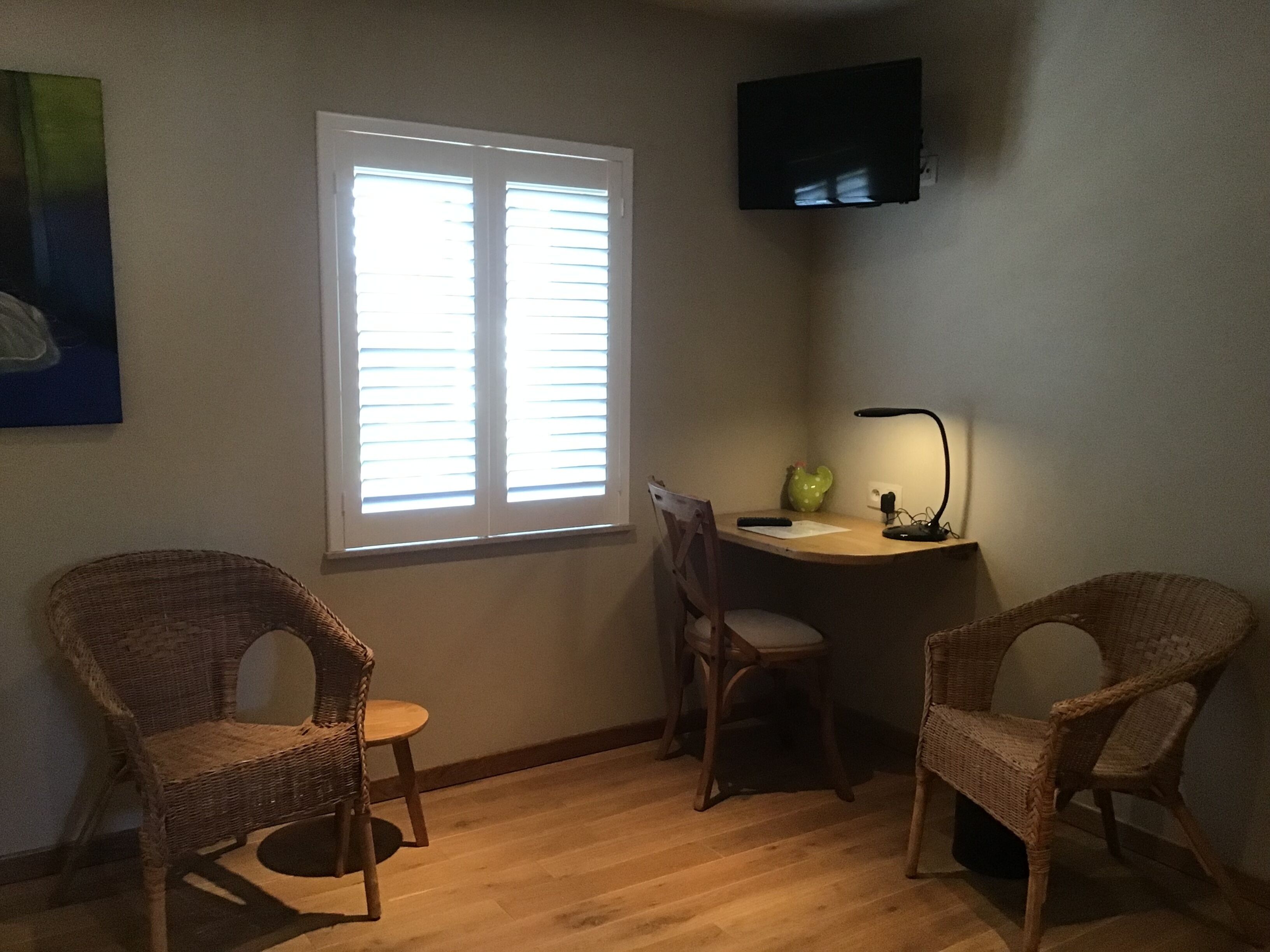 Una habitación con un escritorio de madera, dos sillas de mimbre, un cactus verde, una lámpara y un televisor en la pared.